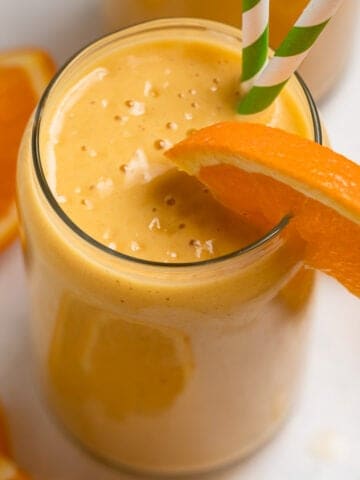 Orange banana smoothie in glass with orange slice to garnish and green striped straws.