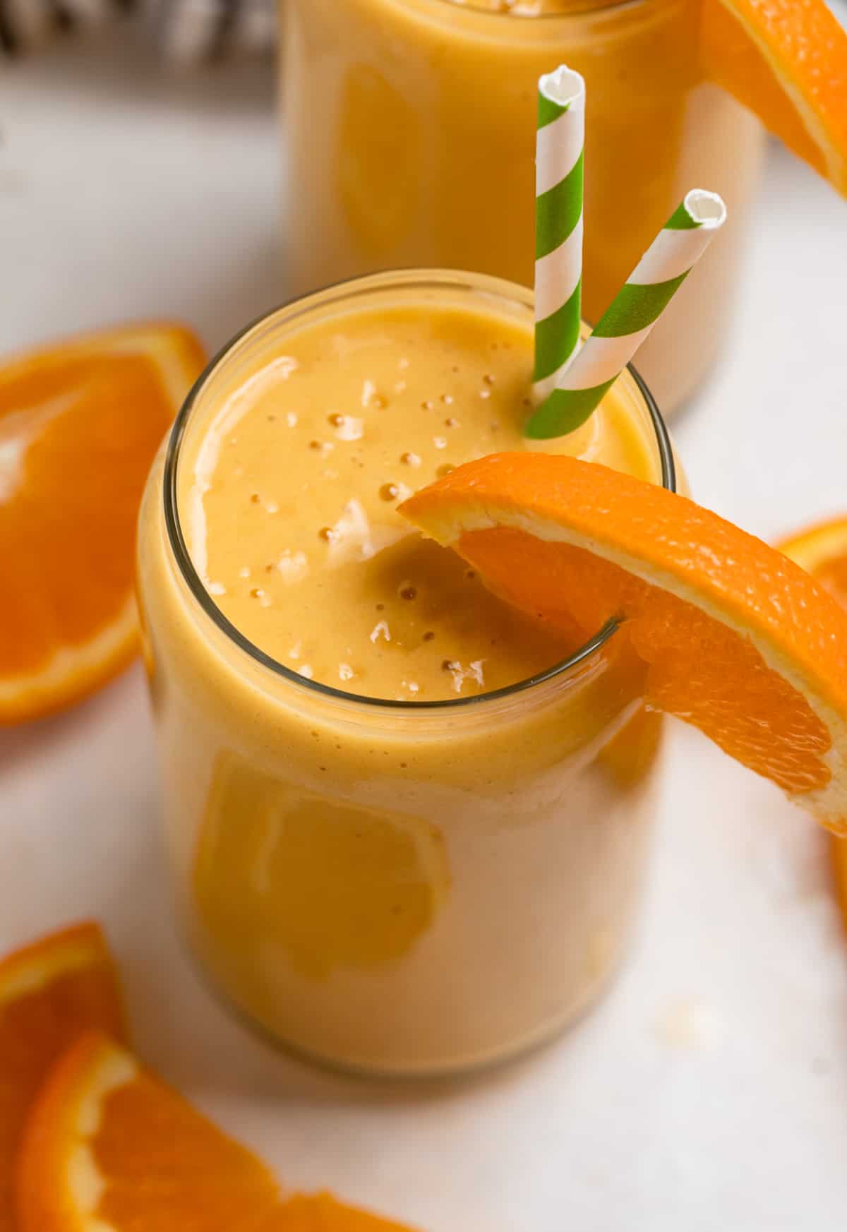 Orange banana smoothie in glass with orange slice garnish and striped straws.