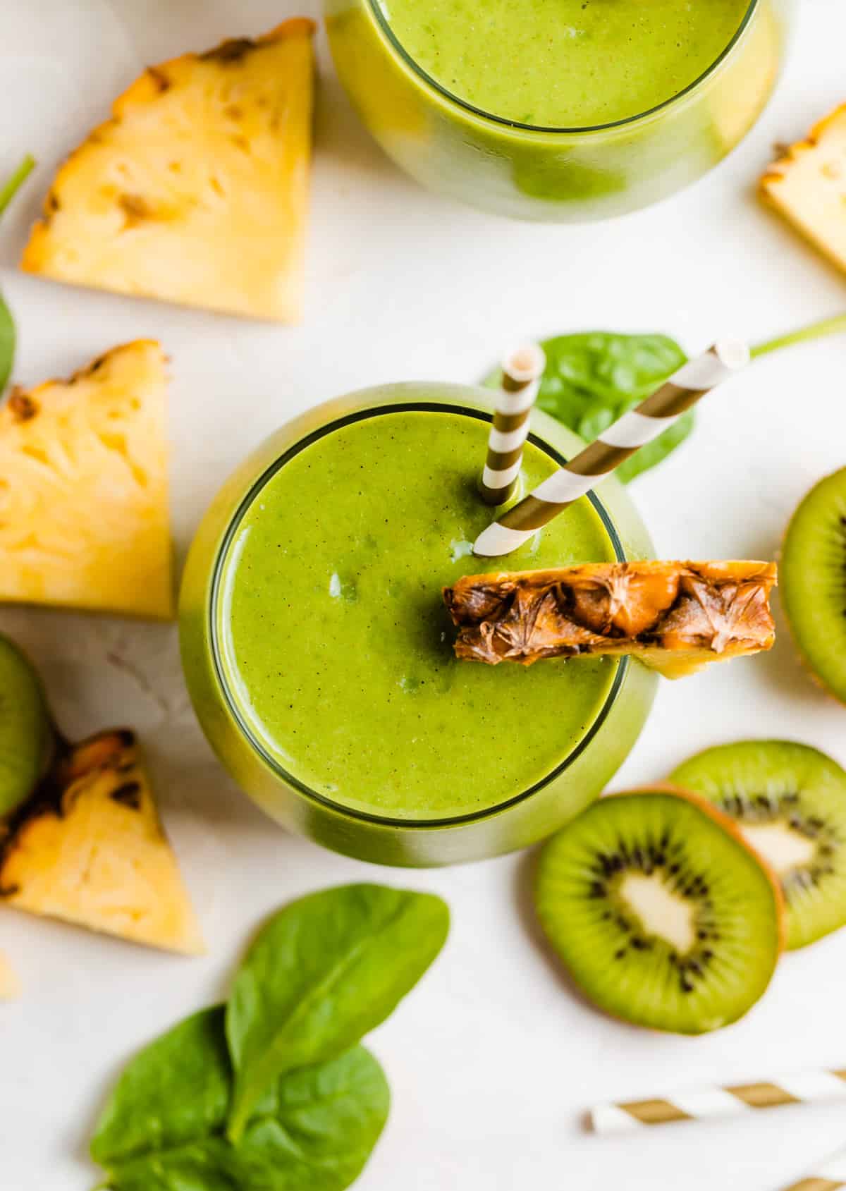 Green kiwi pineapple smoothie in glass with pineapple and kiwi slices surrounding.