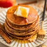 Stack of pancakes on plate with butter and maple syrup and cinnamon sticks beside.
