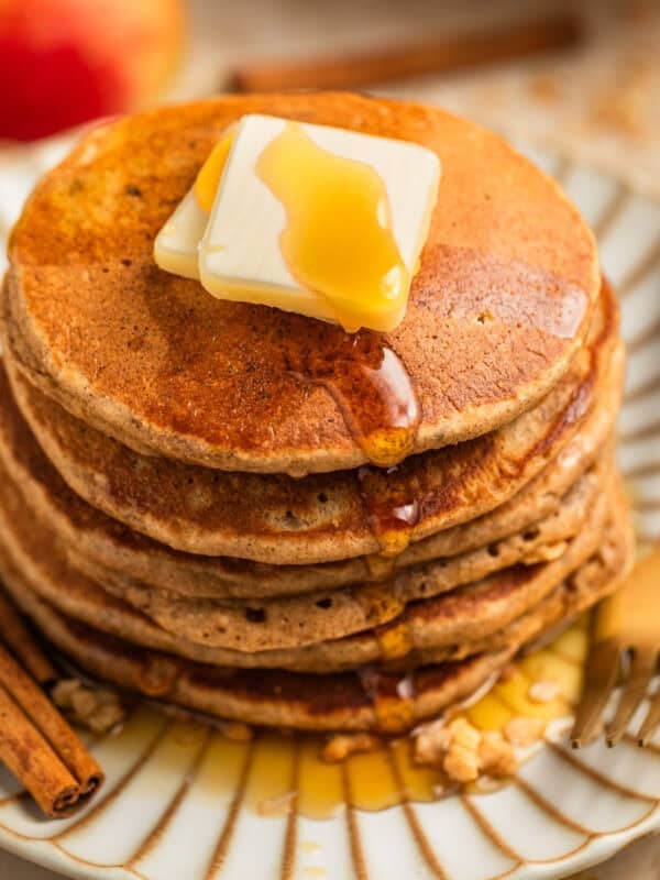 Stack of pancakes on plate with butter and maple syrup and cinnamon sticks beside.