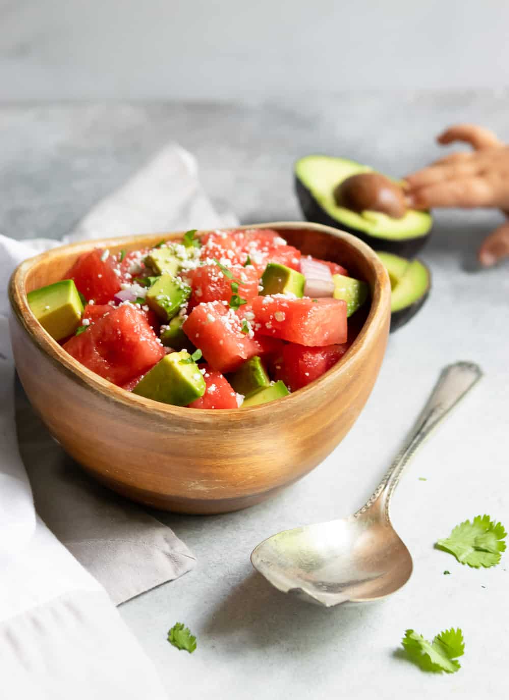 Bowl of watermelon salad with spoon, linen and avocado with child hand reaching for avocado.