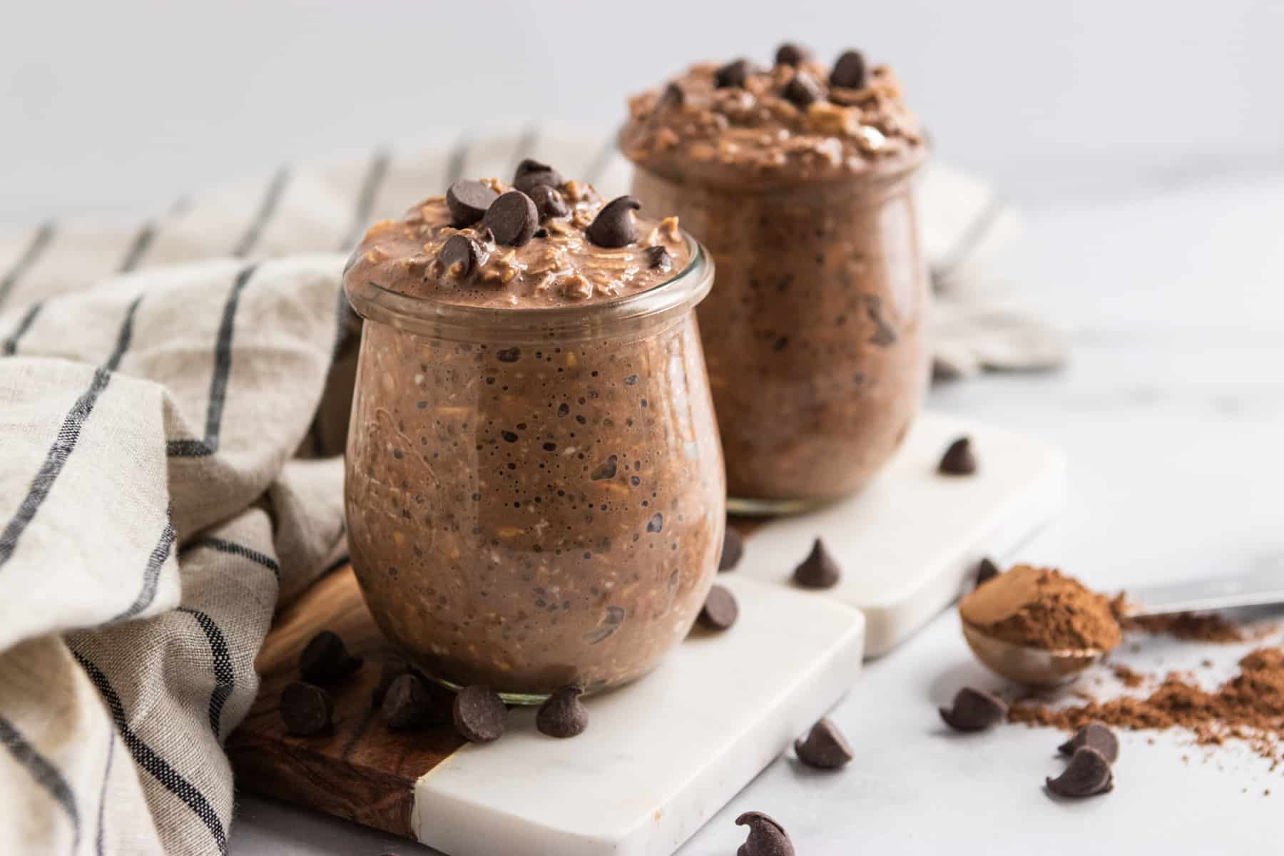 Chocolate Overnight Oats in jars with chocolate chips and coca powder surrounding.