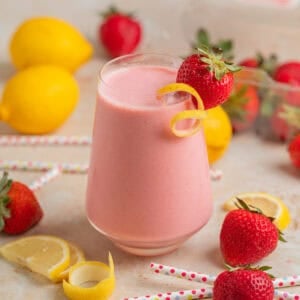 Wine glass with strawberry lemon smoothie with lemon zest and strawberry garnish and berries, lemon slices and straws surrounding.