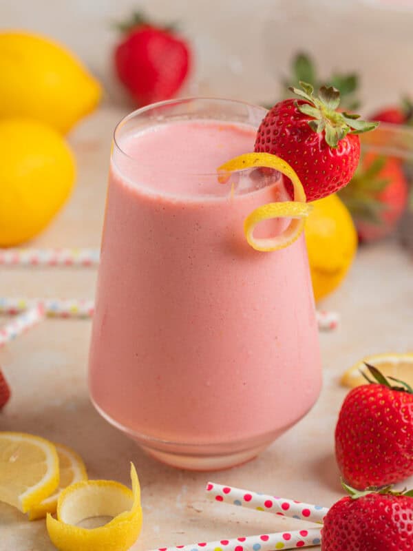 Wine glass with strawberry lemon smoothie with lemon zest and strawberry garnish and berries, lemon slices and straws surrounding.