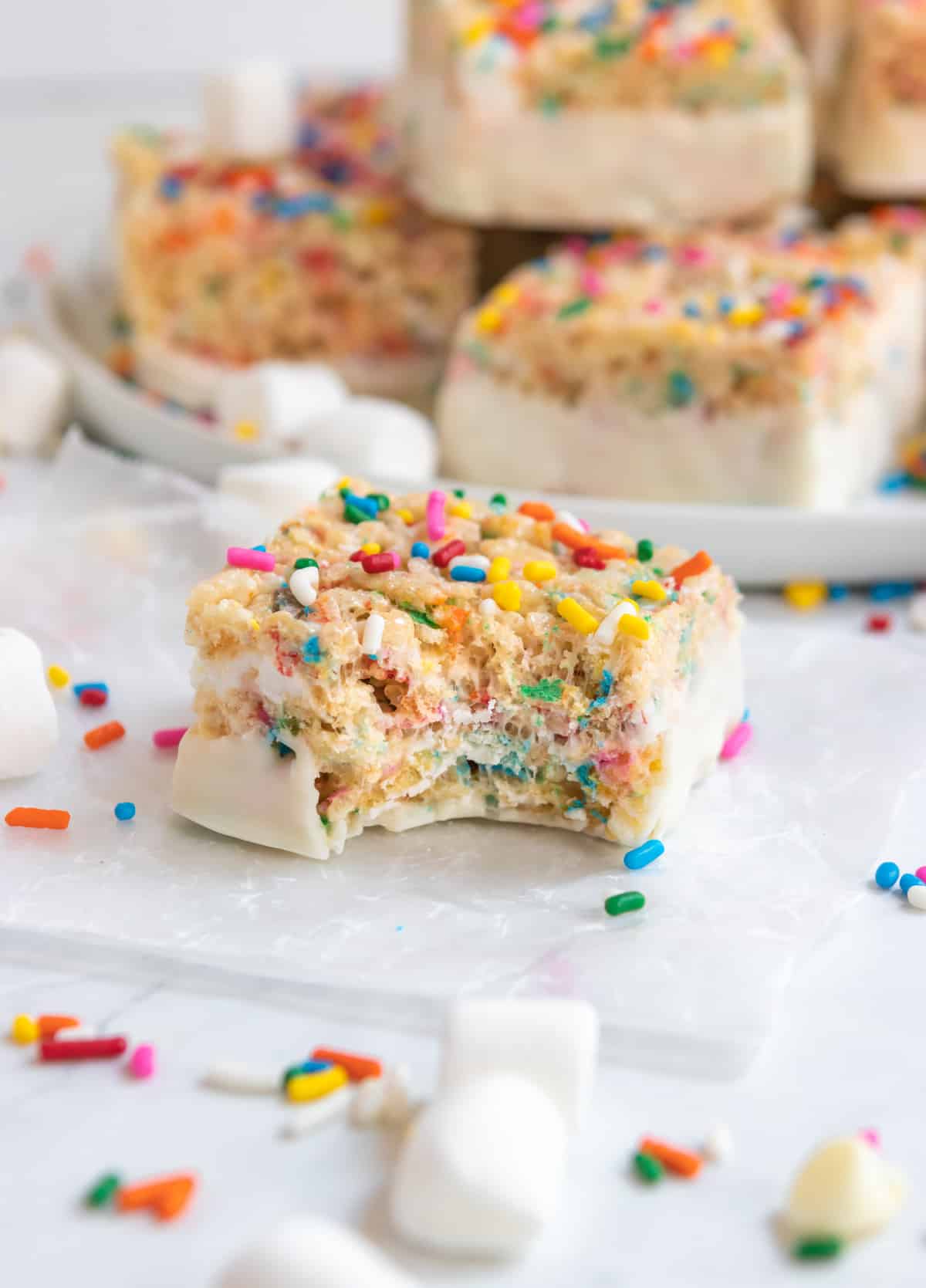 Rice Krispie Treats with sprinkles dipped in white chocolate on parchment.