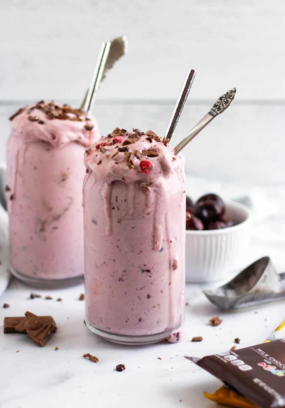 Cherry milkshake in glasses with ice cream scoop and bowl of cherries behind.