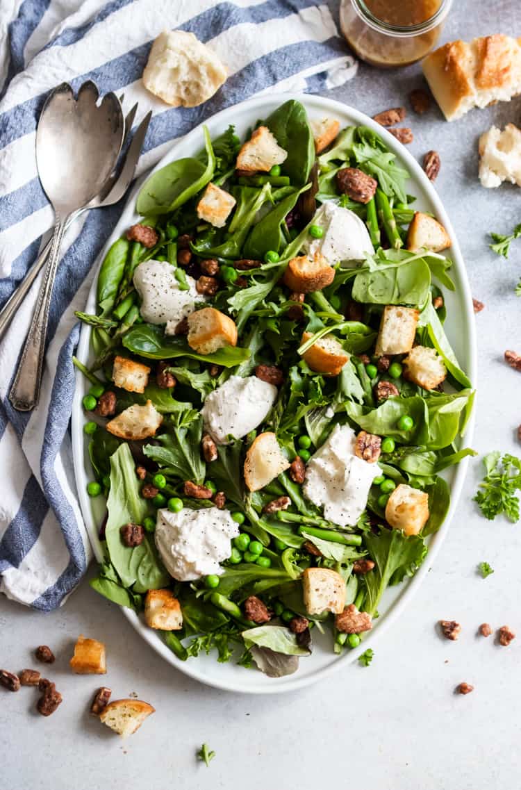 Overhead view of spring green salad with burrata, peas, asparagus and croutons on white serving platter.