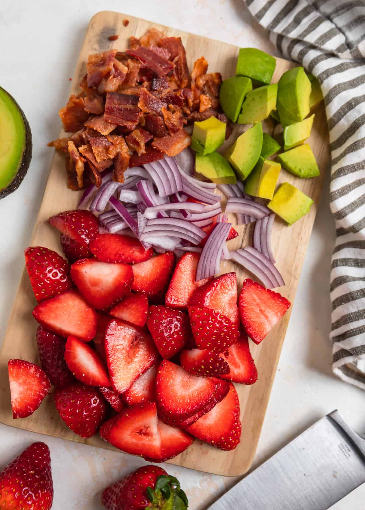 Cutting board with sliced strawberries, avocado, red onion, and bacon.
