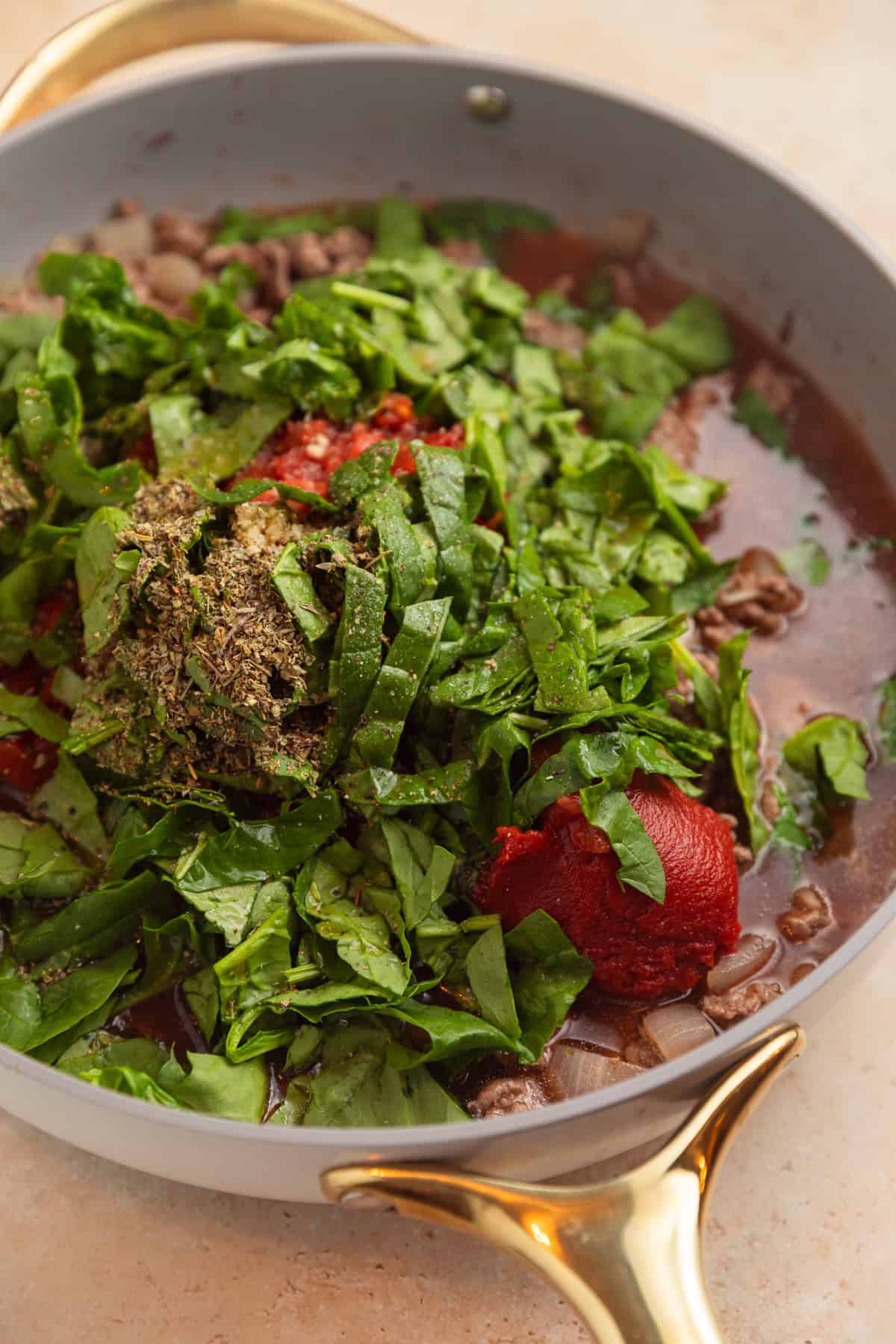Spinach, tomatoes, tomato sauce, and other ingredients added to beef in pan.