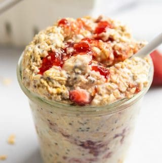 Jar of strawberry cheesecake overnight oatmeal with white spoon and oats on counter surrounding.