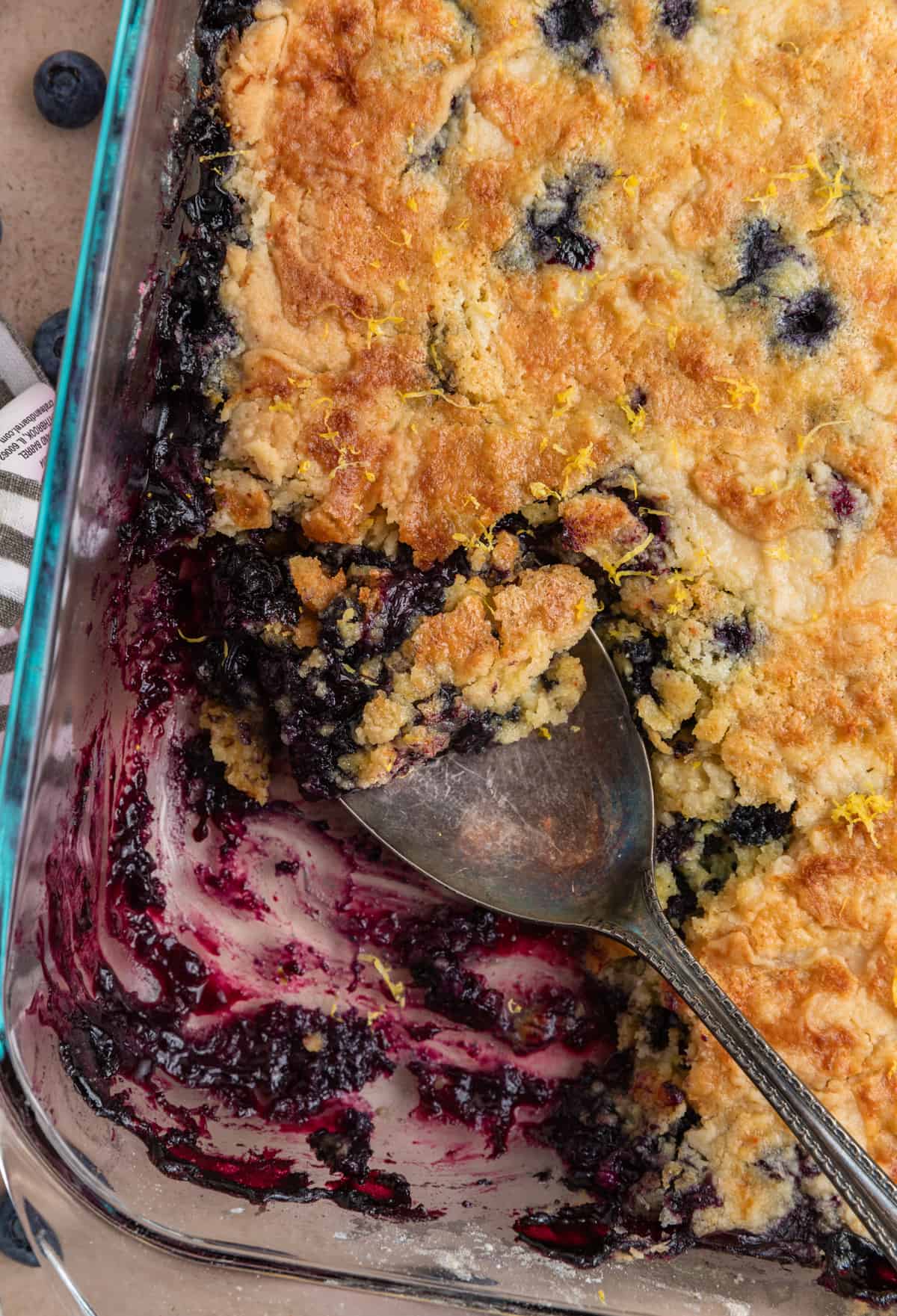Pan with blueberry dump cake and serving spoon scooping up part with lemon zest over top.