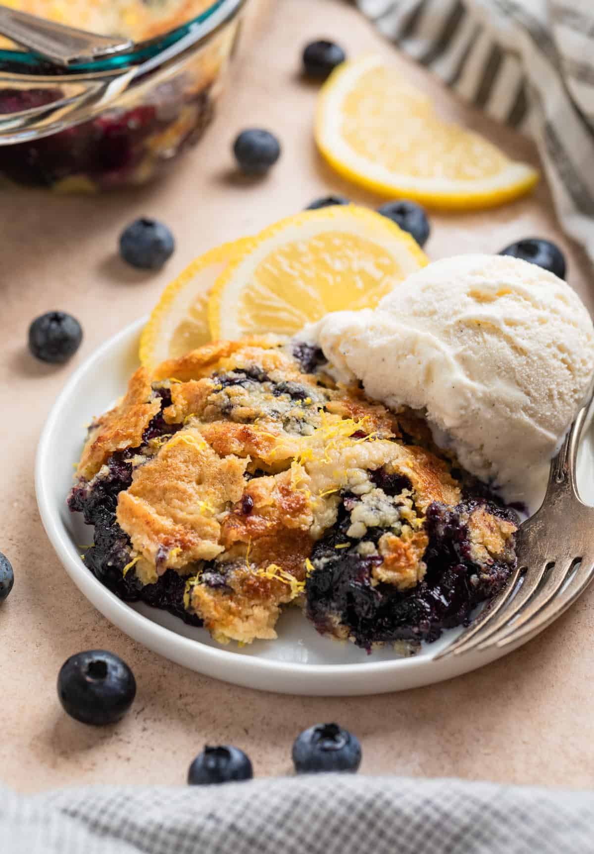 Blueberry lemon dump cake on plate with vanilla ice cream scoop and lemon slices on plate.