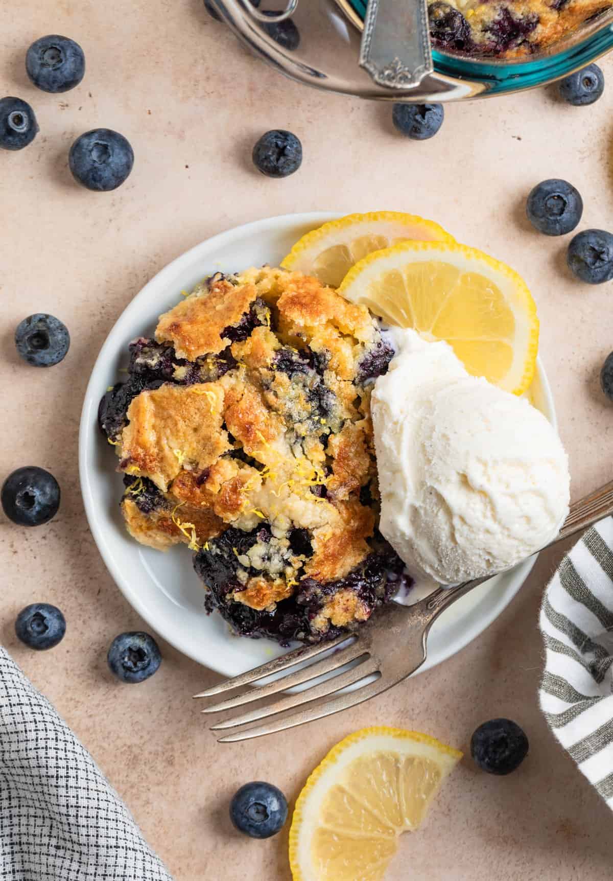 Overhead view of piece of blueberry cobbler on white plate with ice cream and fresh blueberries surrounding.