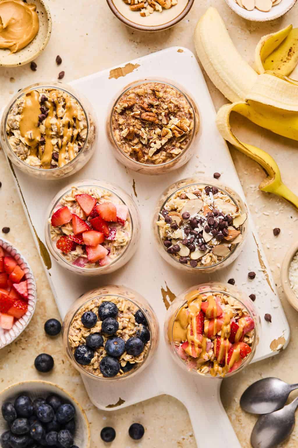 Oatmeal prepared in glasses on white cutting board and topped with berries, chocolate chips, peanut butter, etc.