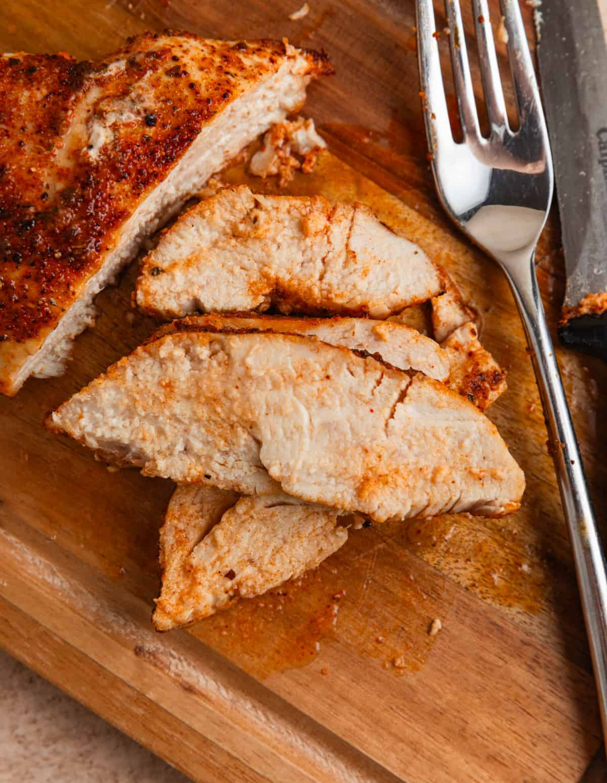 Sliced chicken breast on cutting board with fork and knife.