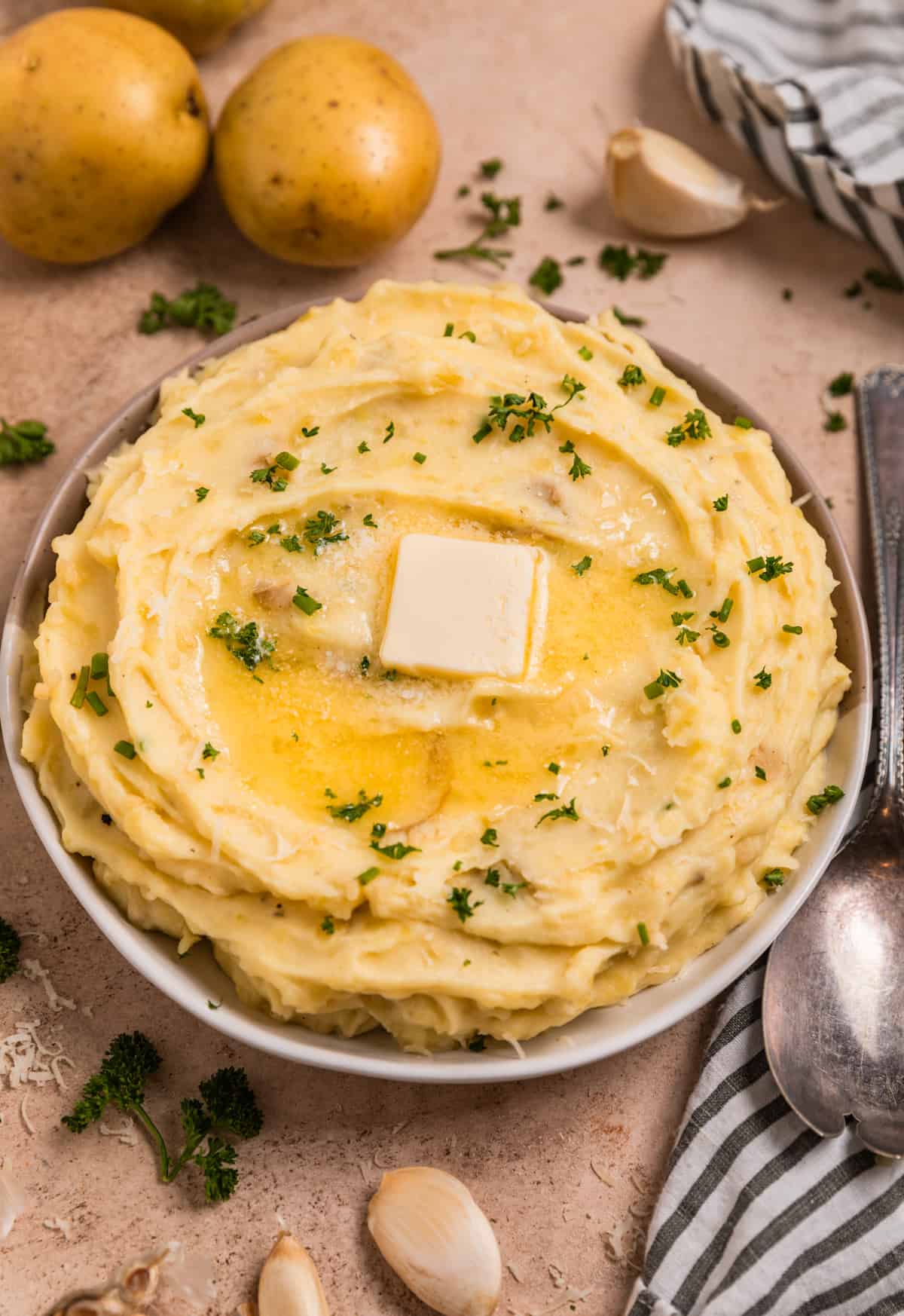 Garlic parmesan mashed potatoes in bowl surrounded by garlic cloves, parsley and other ingredients.