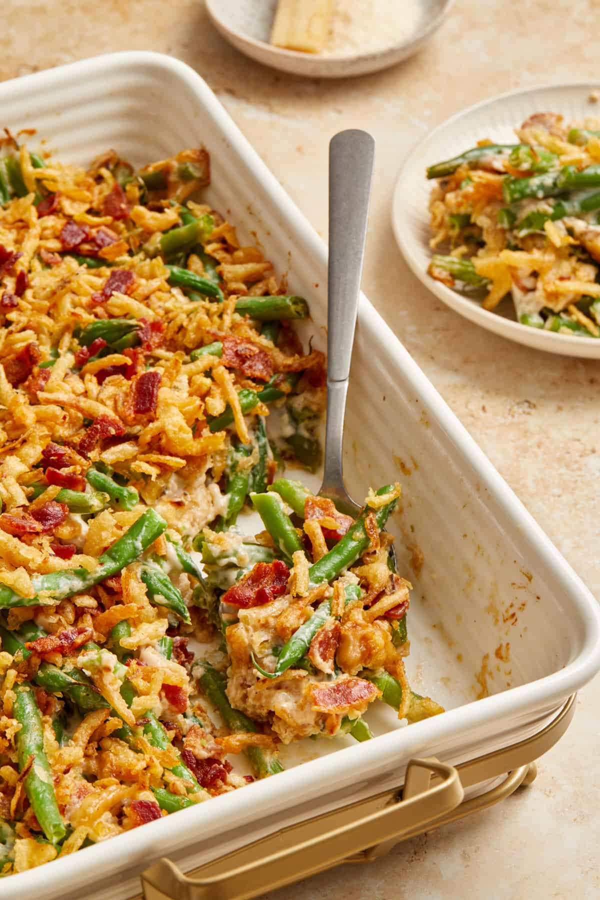 Green bean casserole with chopped bacon in baking pan with serving spoon scooping up a serving.