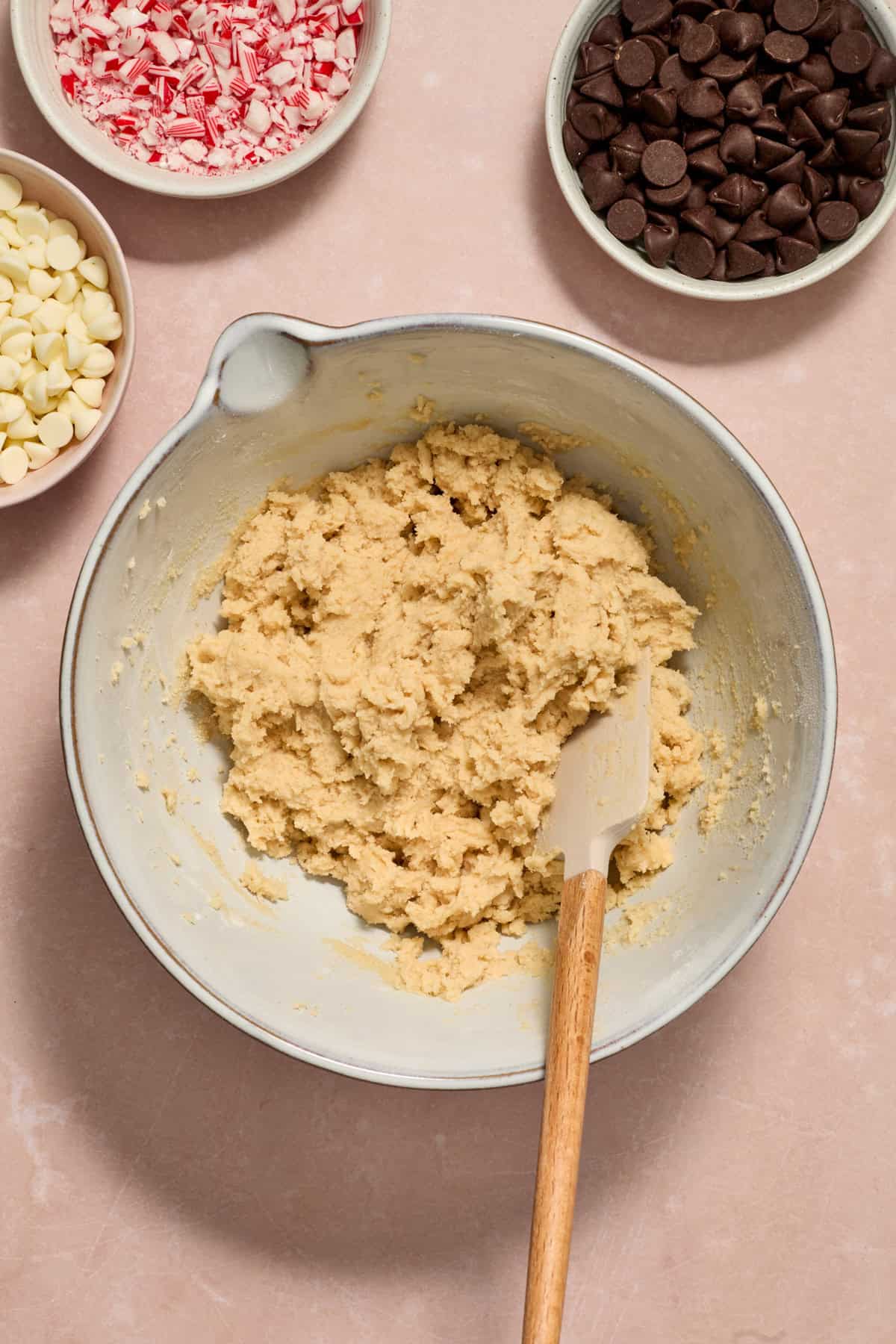 Cookie dough with spatula propped in t and chocolate chips and candy canes in bowls beside.