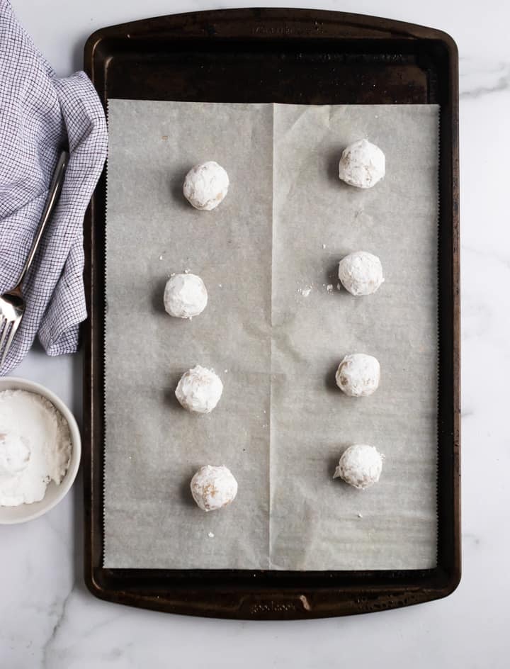 Cream Cheese Snowball Cookies lined up on baking sheet with powdered sugar in bowl beside it.