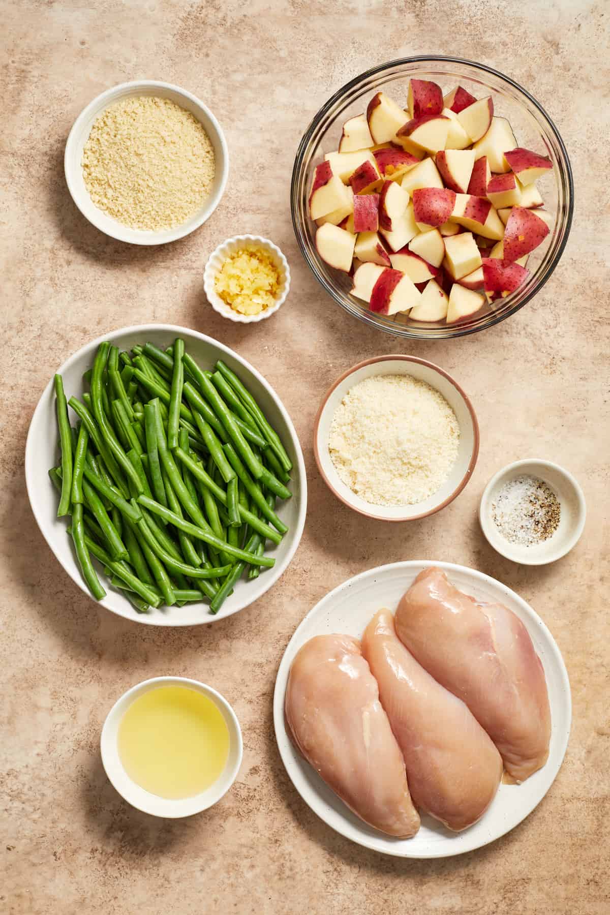 Chicken breast, green beans, potatoes, olive oil, parmesan and other ingredients needed for recipe on surface.