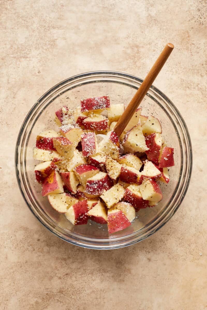 Quartered red potatoes in glass bowl with seasoning and parmesan and spatula.