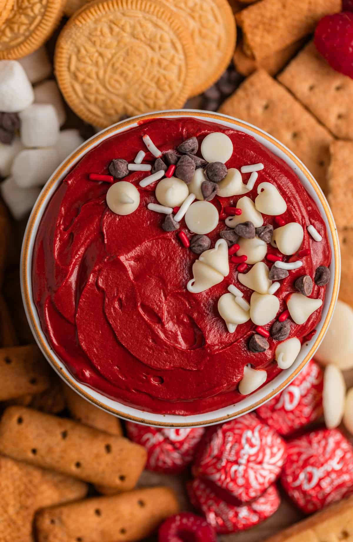 Red velvet cheesecake dip in bowl topped with sprinkles and chocolate chips surrounded by cookies, graham crackers and other dippers.
