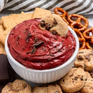 Red velvet cheesecake dip in white bowl with chocolate chip cookie.