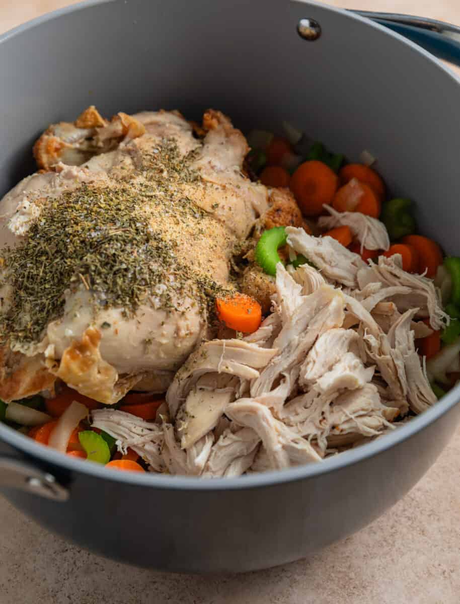 Shredded rotisserie chicken and chicken carcass in pot with carrots, celery and onion.