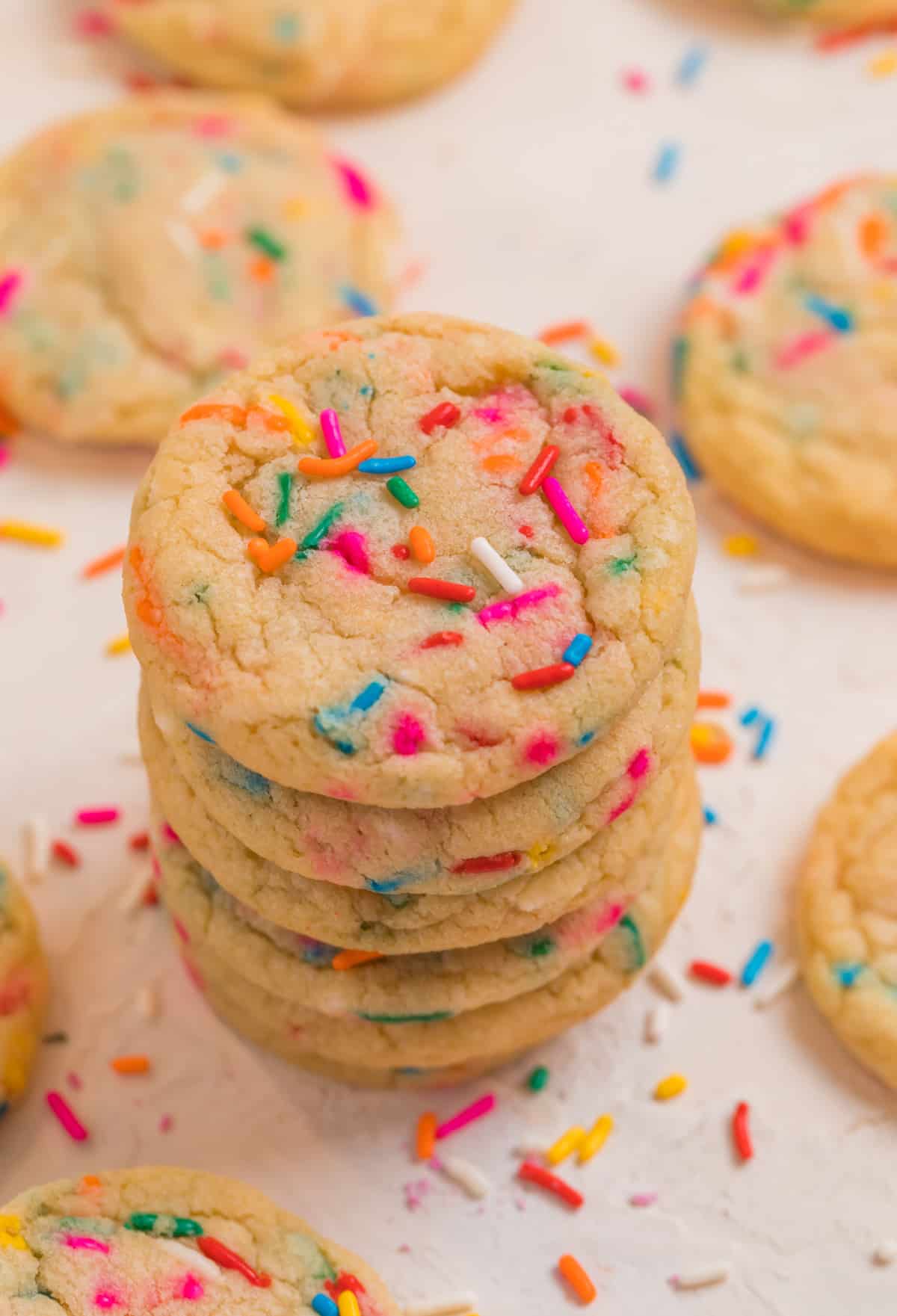 Stack of birthday cake cookies with other cookies and sprinkles surrounding the cookie stack.