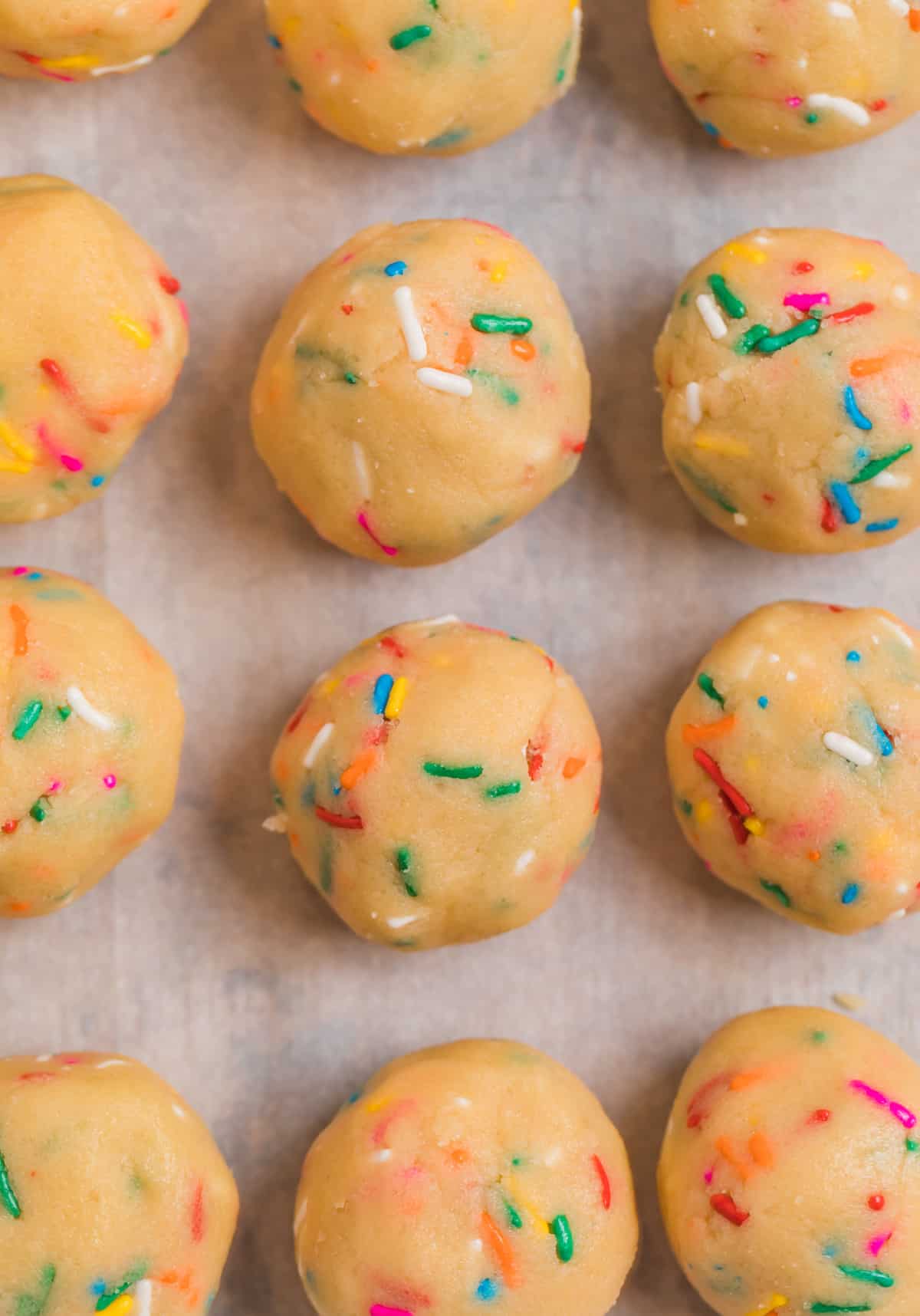 Rolled cookie dough balls with rainbow sprinkles on parchment lined baking sheet.