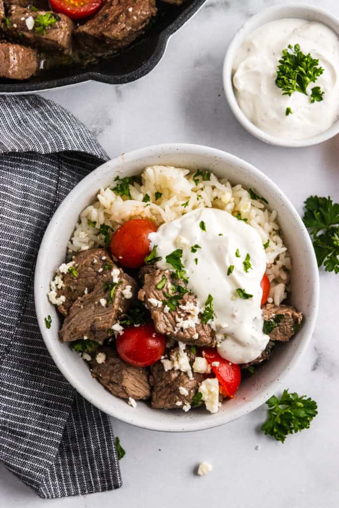 Overhead shot of bowl with steak bites, tomatoes, parsley, feta and yogurt sauce.