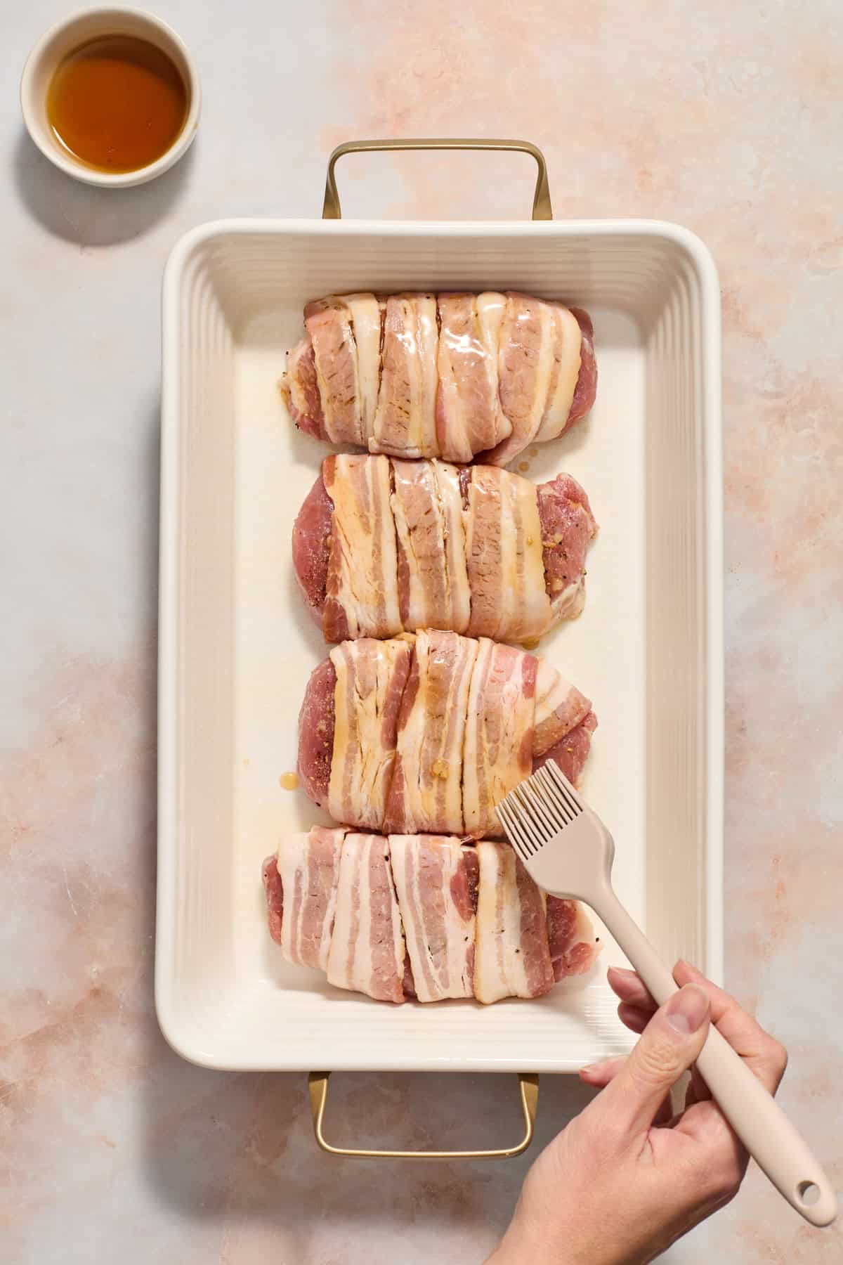 Maple being brushed on bacon wrapped pork chops in baking dish with dish of maple syrup beside the pan.