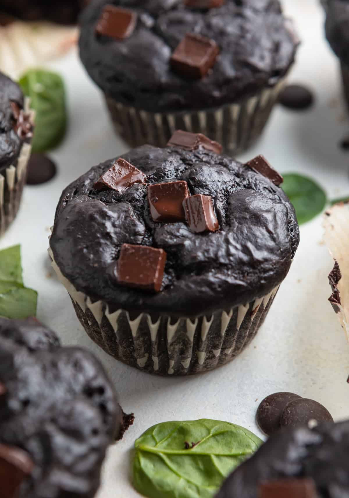 Chocolate spinach muffins on counter with chocolate chips and spinach surrounding.