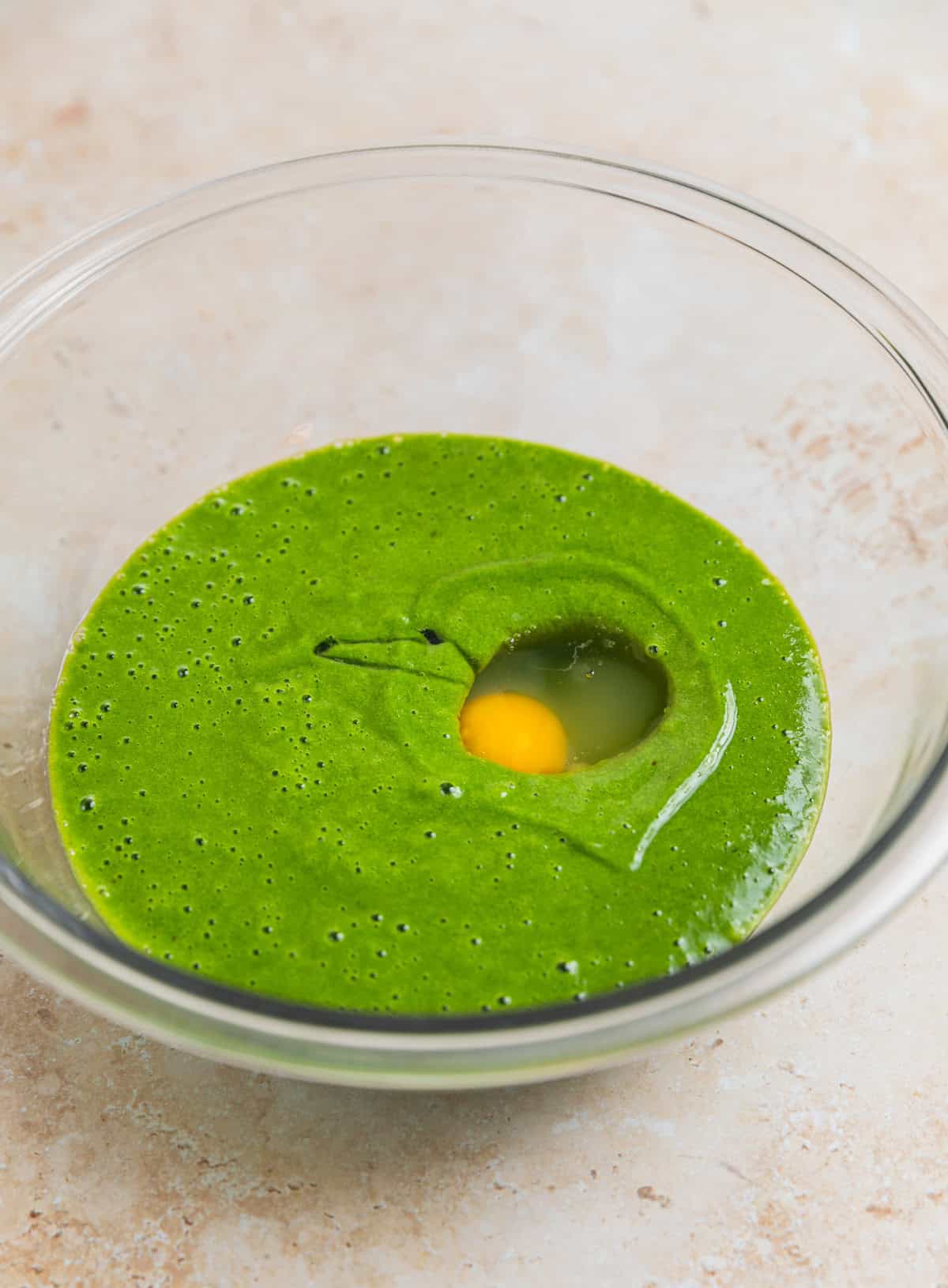 Glass mixing bowl with spinach and banana mixture and egg added to it.