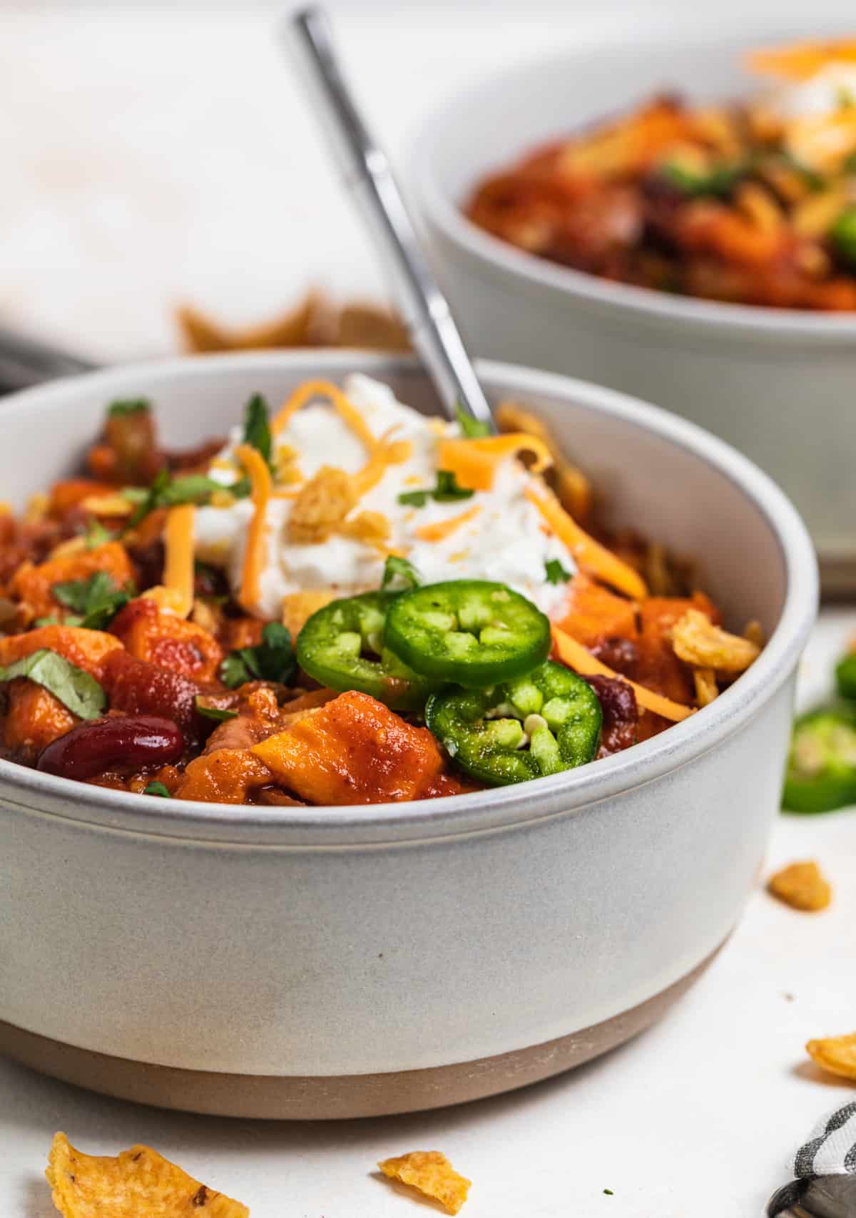 Bowls with slow cooker sweet potato veggie chili topped with cheese, jalapeño, corn chips and spoon inside bowl.