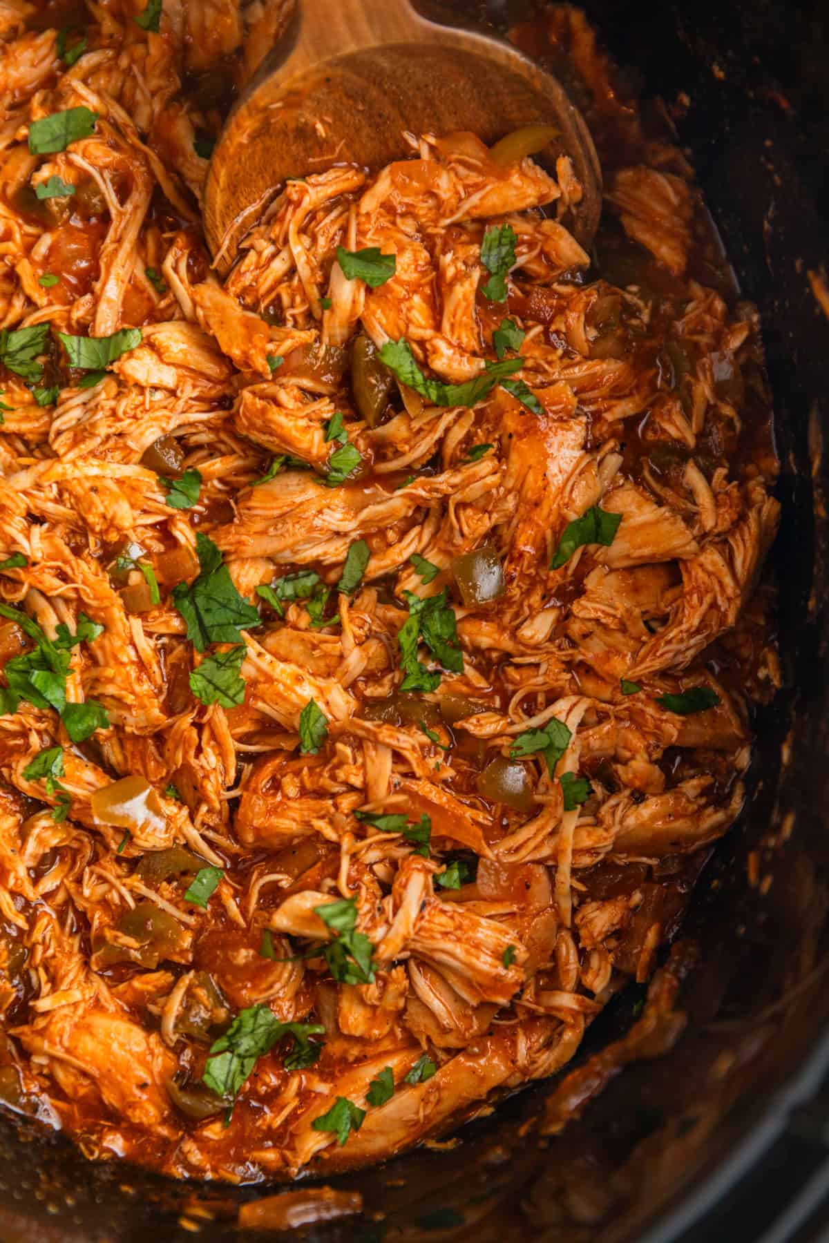 Shredded bbq chicken topped with chopped cilantro in crockpot with wooden spoon scooping in.