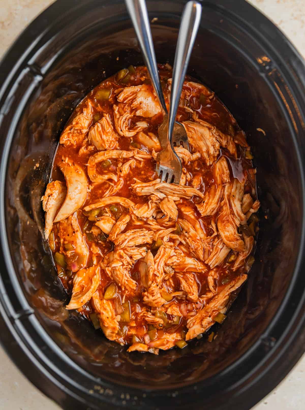 Forks shredding bbq chicken breast in slow cooker pot.