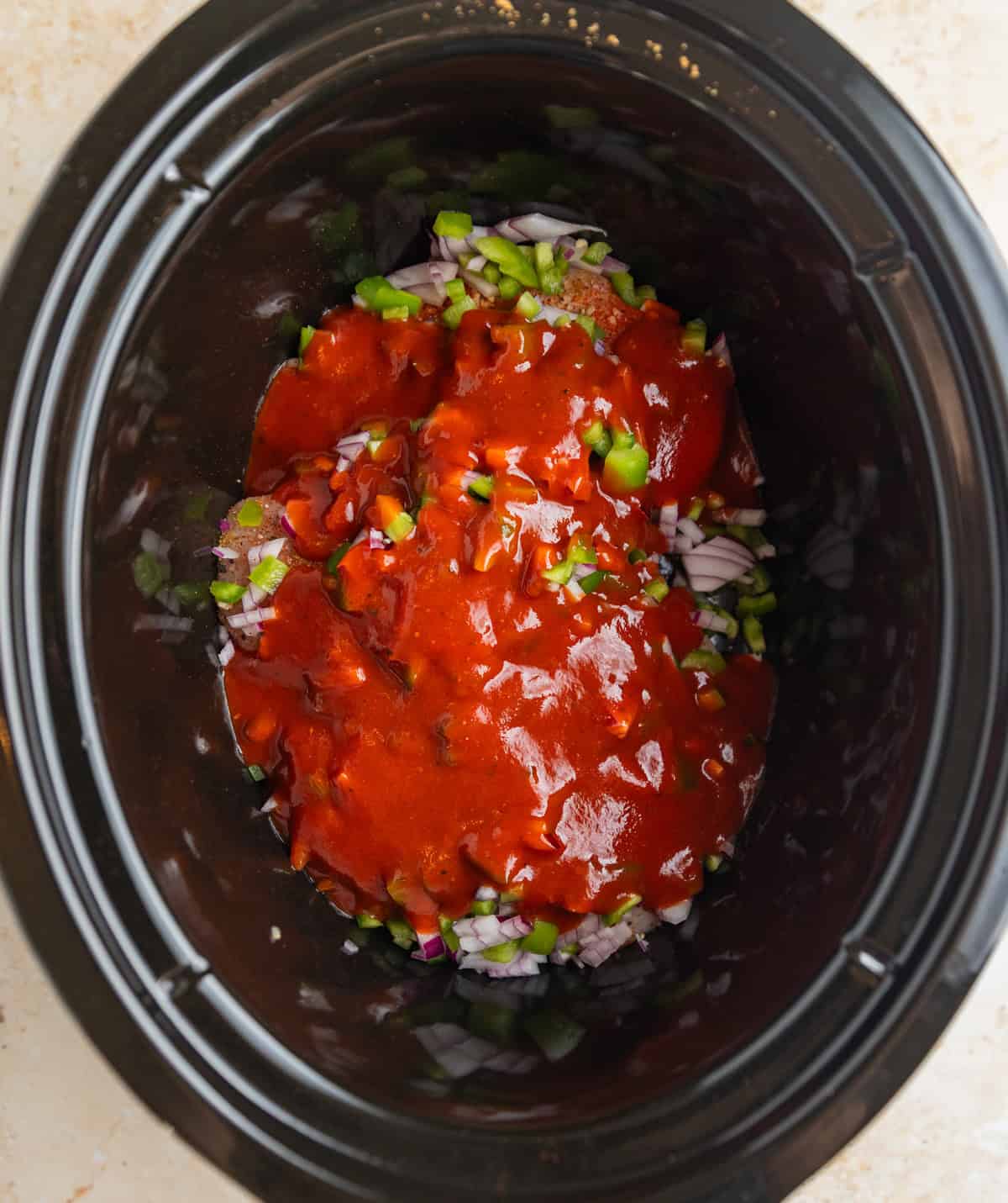 Chopped pepper and onions over seasoned chicken breast topped with barbecue sauce in crockpot before cooking.