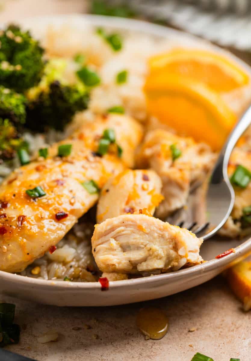 Piece of baked orange chicken on fork with plate of broccoli and rice.