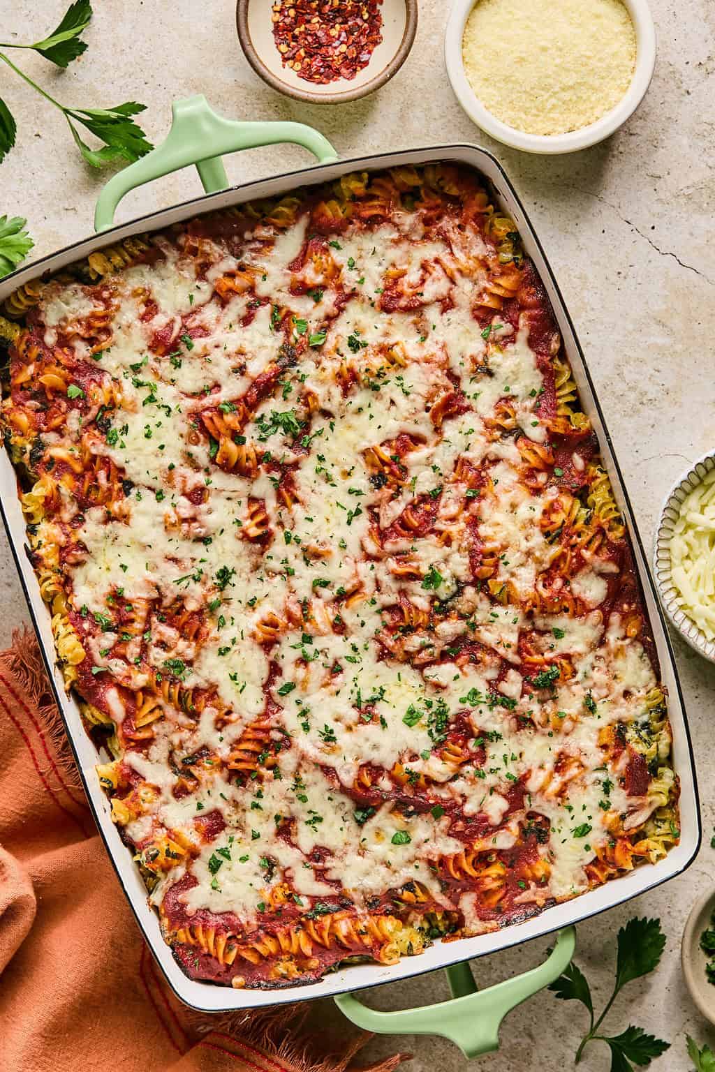 Baked pasta topped with fresh herbs and bowl of spices and cheese surrounding the dish.