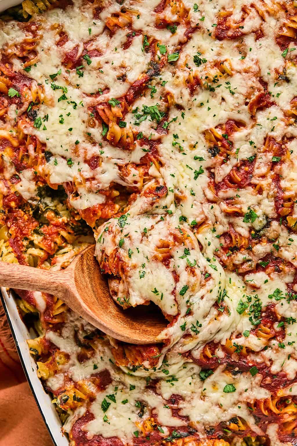 Wooden spoon scooping into baked pasta with spinach topped with chopped parsley.