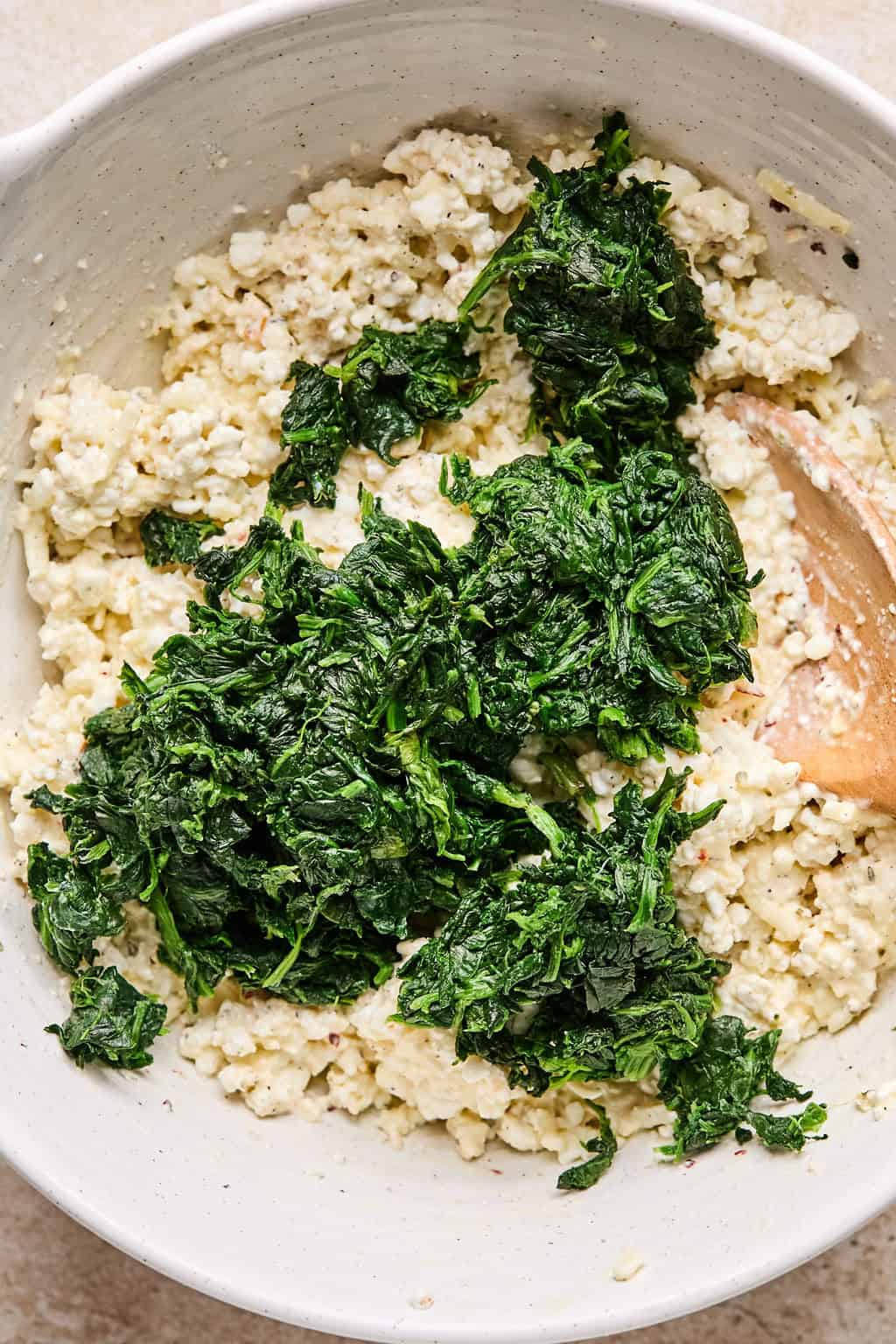 Spinach added to cottage cheese mixture in mixing bowl with wooden spoon.