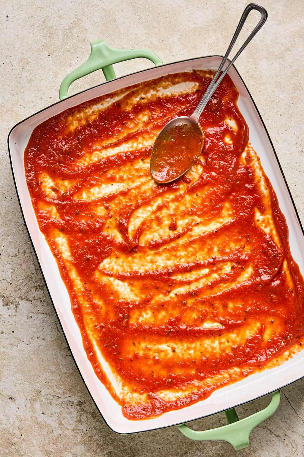 Marinara sauce spread in the bottom of a 9x13 inch pan with spoon.
