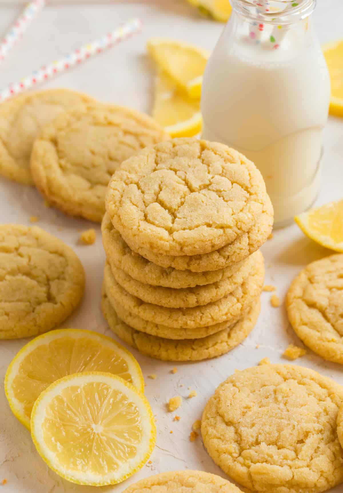 Lemon sugar cookies stacked on top of each other with glass of milk behind and other cookies and lemons surrounding.