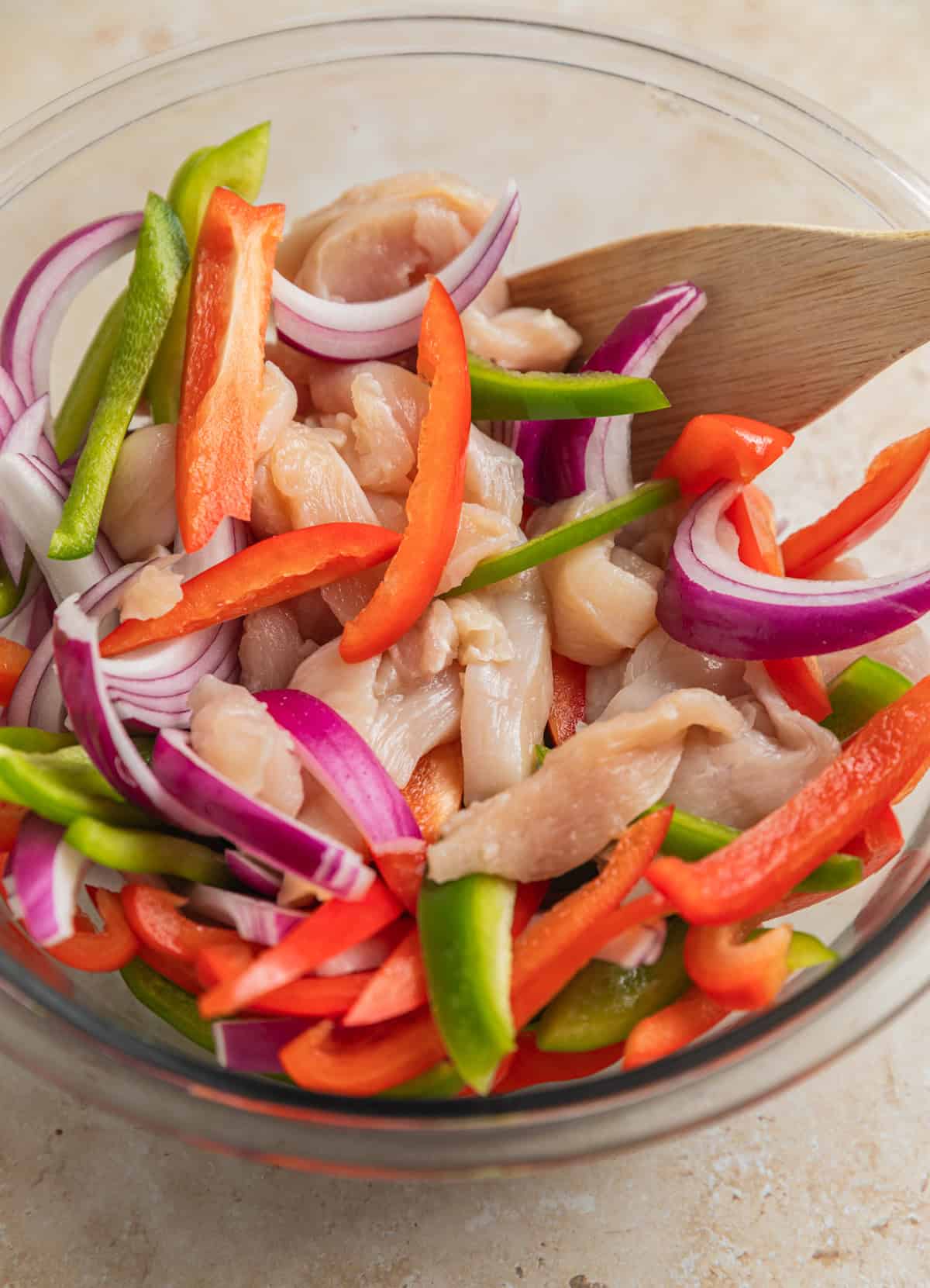 Raw sliced chicken, sliced red and green bell peppers and red onion in glass bowl with wood spoon.