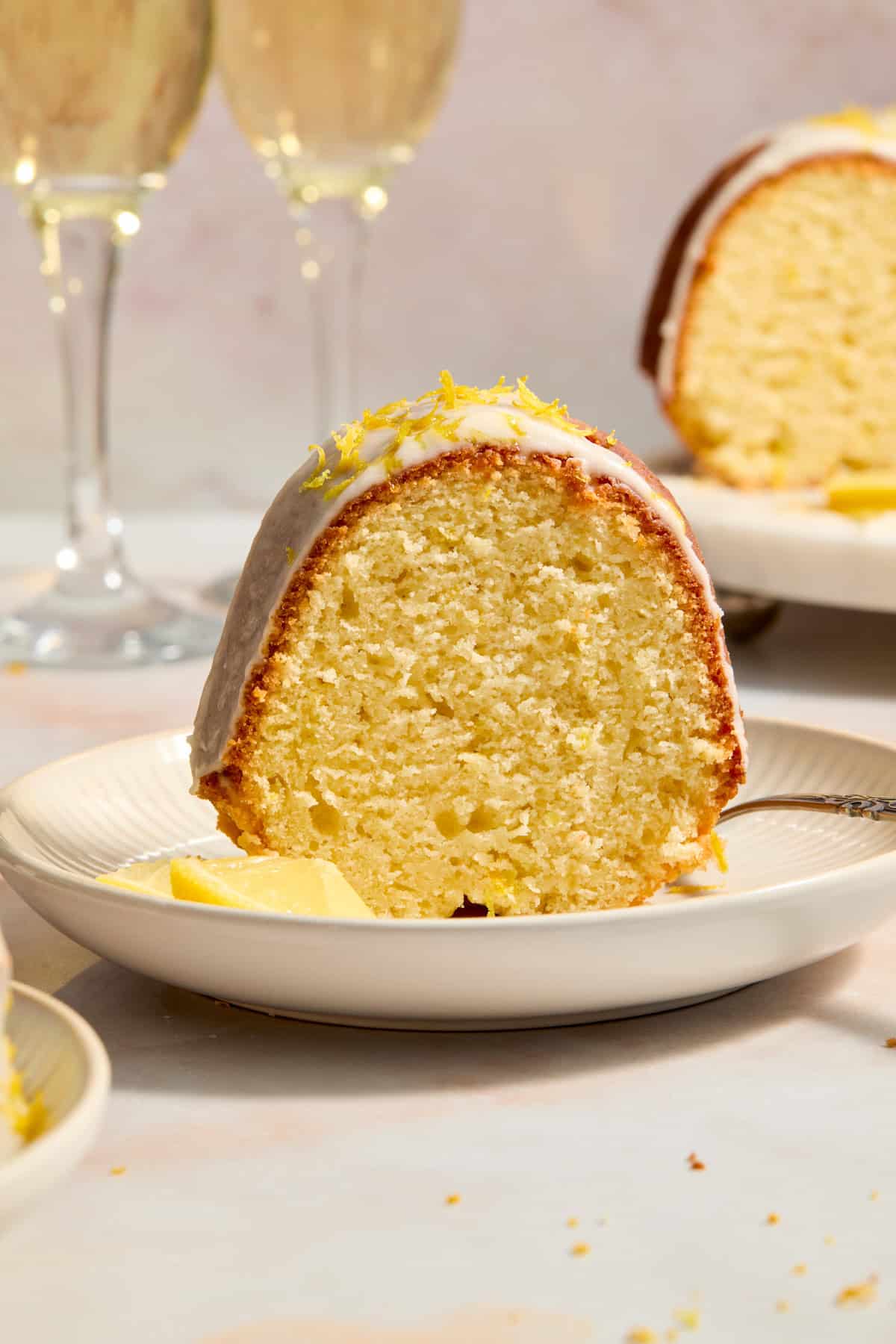 Slice of lemon bundt cake on plate with lemon slices and glasses of champagne in the background.