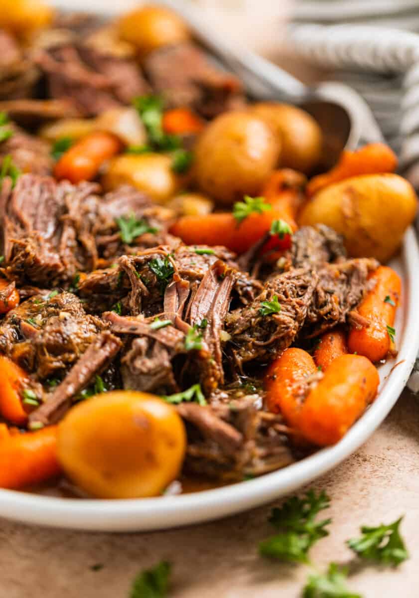 Crockpot chuck roast on white serving platter with carrots, onion and petite potatoes and topped with chopped parsley.
