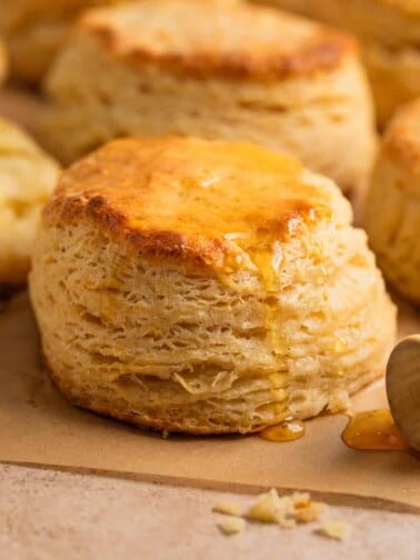 Simple honey biscuits on tan parchment with honey drizzling over top with honey dipper stick beside it.