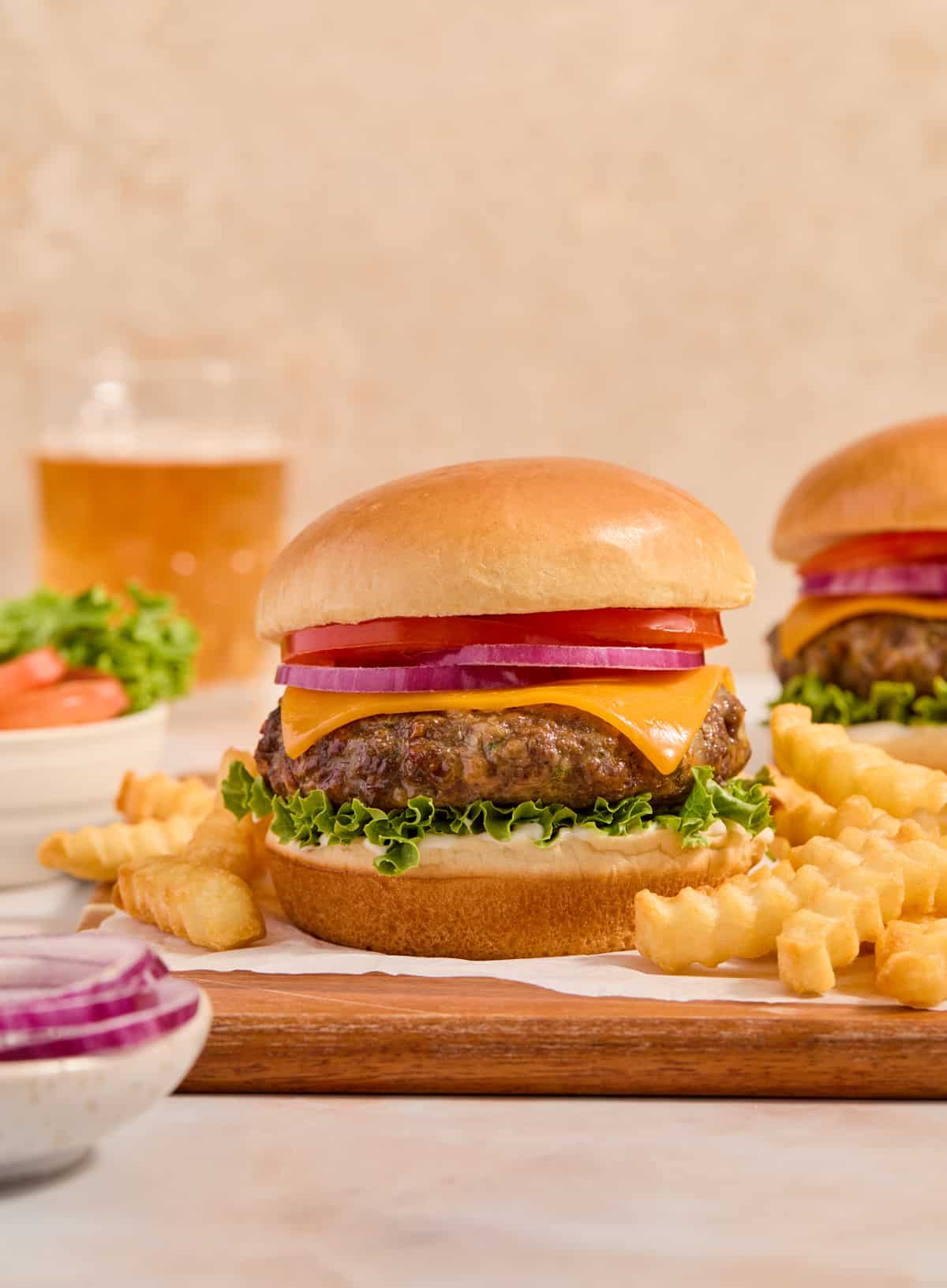 Burger on bun with cheese, lettuce, onion and tomato with beer glass in the background and fries on the platter.